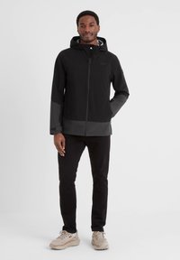 Black waterproof jacket with grey accents, zip front, and hood. Features a smooth texture and minimalist design, worn over black pants and sneakers.