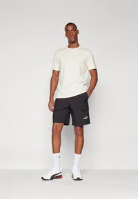 Άνδρας που στέκεται φορώντας κρέμ μπλούζα, μαύρες σορτς Puma με πλαϊνές τσέπες, άσπρες κάλτσες και άσπρα αθλητικά παπούτσια με μαύρες και κόκκινες λεπτομέρειες.