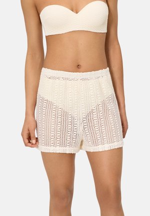 Mujer vistiendo pantalones cortos de ganchillo blanco de talle alto y top bandeau sin tirantes a juego con textura, sobre un fondo liso.