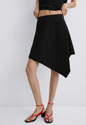 Mujer que lleva una falda negra asimétrica y sandalias rojas de tacón, de pie y ajustándose la cintura de la falda con las manos.