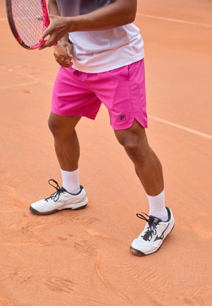 Roze sportshorts met zijslitsen, logo aan de linkerkant, gecombineerd met witte sportschoenen en witte sokken, op een gravel tennisbaan.