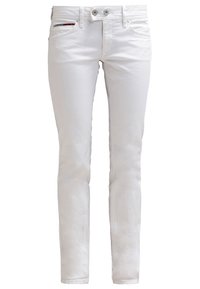 Białe, dopasowane jeansy z dwoma przednimi guzikami, suwakiem, przednimi kieszeniami oraz małym dekoracyjnym paskiem w kolorach czerwonym, białym i czarnym na prawej kieszeni.