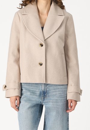 Vrouw draagt een beige cropped wollen jas met grote revers en met knopen gesloten manchetten over lichtblauwe jeans.