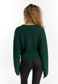 Maglione verde lavorato a maglia con una vestibilità ampia, caratterizzato da maniche larghe e orlo a coste. Indossato con pantaloni neri aderenti, che evidenziano il contrasto di texture e colore.