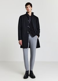 Schwarzer Mantel mit hohem Kragen und Knöpfen, kombiniert mit einem dunklen Hemd und hellgrauen, zulaufenden Hosen. Das Model trägt schwarze Stiefeletten.