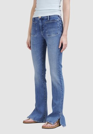 Person trägt blaue Jeans mit aufgesetzten Fronttaschen und Seitenschlitzen am Saum, kombiniert mit offenen Sandalen und einem hellblauen Top.