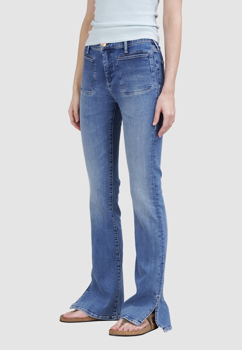 Personne portant un jean bleu avec des poches plaquées à l'avant et des fentes latérales à l'ourlet, associé à des sandales ouvertes et un haut bleu clair.