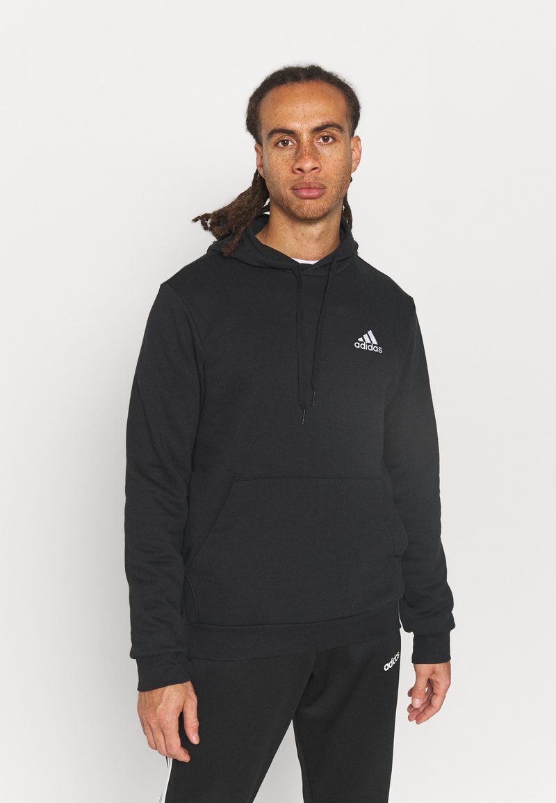 Sudadera negra de tela suave, con un bolsillo frontal, capucha con cordón y un pequeño logo blanco de Adidas en el pecho.