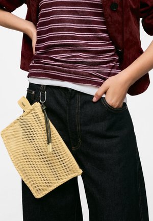 Person trägt ein bordeauxfarbenes gestreiftes Oberteil und dunkle Jeans mit einer beigen Netz-Clutch, die an der Gürtelschlaufe befestigt ist.