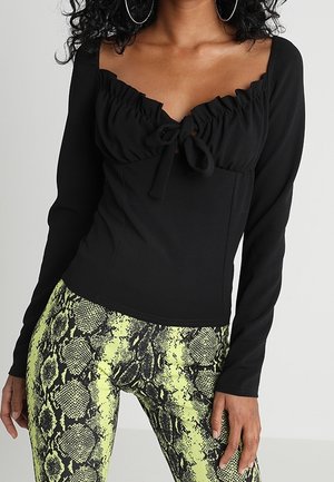 Femme portant un haut noir à manches longues froncé avec un lien devant et un legging vert à motif serpent, debout devant un fond clair uni.