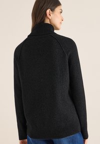 Schwarzer, gerippter Rollkragenpullover mit weicher Textur und lockerer Passform, mit Raglanärmeln und hohen Kragen, von hinten gezeigt.