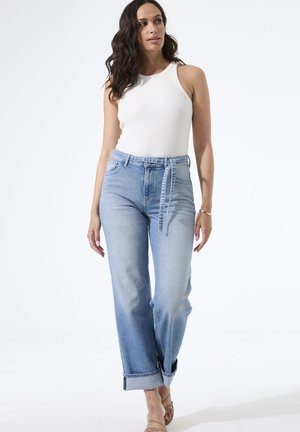 Femme aux longs cheveux foncés portant un haut blanc sans manches, un jean taille haute bleu clair avec ceinture, et des sandales ouvertes beige, debout.