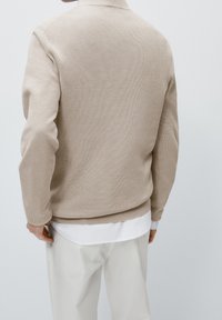 Person, die einen beigefarbenen Strickpullover über einem weißen Hemd und einer weißen Hose trägt, von hinten vor einem schlichten hellen Hintergrund gezeigt.