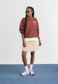 Sweatshirt castanho com logótipo branco, por cima de uma camisa branca, combinado com uma saia de ganga bege. Complementado com sapatilhas rosas e óculos de sol.