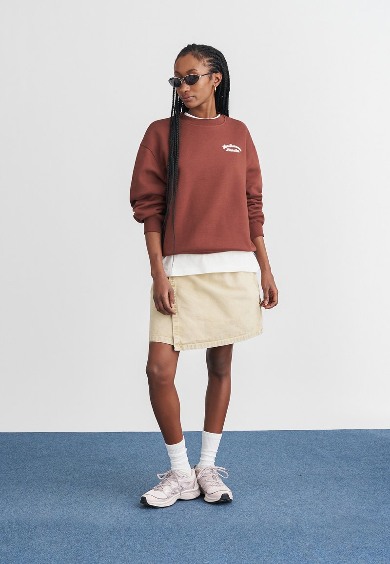Brauner Sweatshirt mit weißem Logo, über einem weißen Shirt getragen, kombiniert mit einem beigen Jeansrock. Abgerundet mit rosa Sneakers und Sonnenbrille.