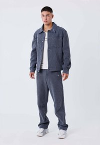 Giacca a quadri blu navy con chiusura a bottoni, dotata di due tasche frontali, abbinata a pantaloni coordinati e sneakers bianche. Tessuto semplice e strutturato.