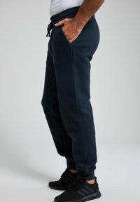 Dunkelblaue Sweatpants mit einer Kordelzugtaillierung, seitlichen Taschen, gerippten Bündchen und einer weichen Textur. Getragen mit schwarzen Sportschuhen.