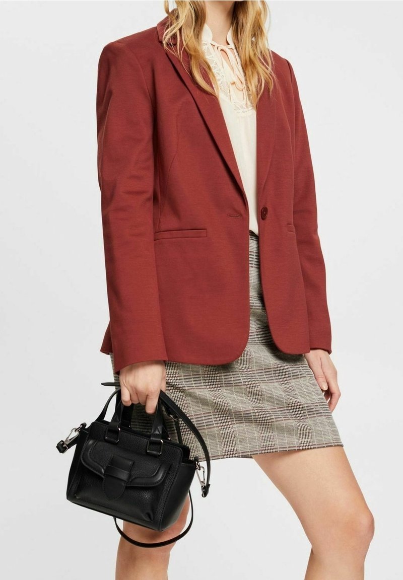 Femme portant un blazer rouille, une blouse crème, une jupe à carreaux, tenant un sac à main en cuir noir à ses côtés sur fond blanc.