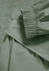 Světle zelená látka se žebrovanou strukturou, elastickými manžetami a vrstveným designem. Hladký povrch s pečlivými detaily švů.