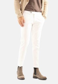 Persoon in witte slim-fit broek, beige textuurcardigan, bruine shirt en taupe suède enkellaarsjes staand tegen een effen achtergrond.