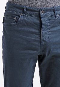 Blauwe katoenbroek met een gladde textuur, voorzien van een rits, knoopsluiting en voorzakken. Het ontwerp bevat decoratieve stiksels.