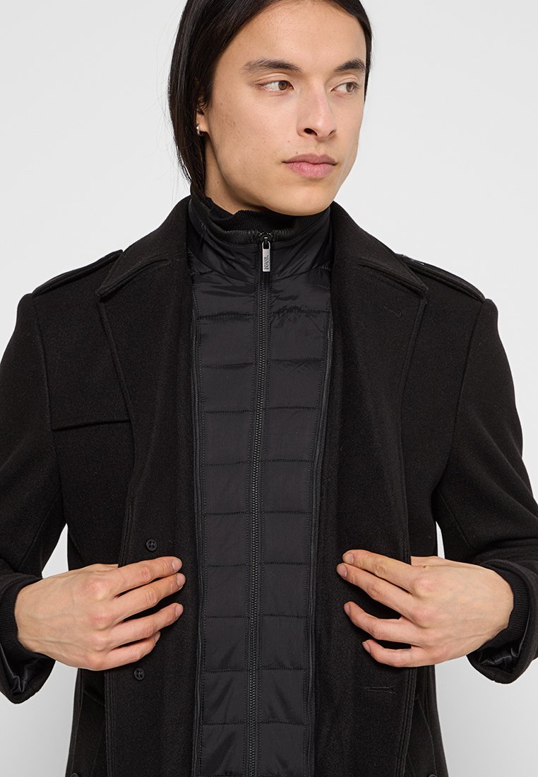 Schwarzer Wollmantel über einer schwarzen gesteppte Innenjacke geschichtet. Merkmale sind ein Stehkragen, Knöpfe vorne und Klappentaschen. Glatte Textur.