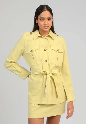 Femme aux longs cheveux noirs portant une veste ceinturée jaune pâle avec poches et boutons, assortie à une jupe, posant devant un fond gris.