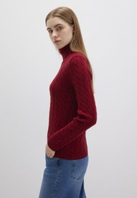 Kabelstrick-Pullover mit Rollkragen in Dunkelrot, mit strukturierten Mustern und enganliegenden langen Ärmeln, kombiniert mit hellblauen Jeans.