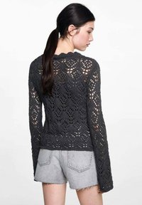 Schwarzer, aus Spitzenstrick gefertigter Pullover mit langen Ärmeln, gewelltem Ausschnitt und aufwendigen Diamanten- und Blumenmustern, kombiniert mit grauen Jeans-Shorts.