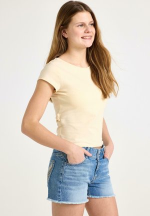 Ung kvinde smilende, iført en lysegul kortærmet top og blå denimshorts, med hænderne i forlommerne, kiggende mod højre.