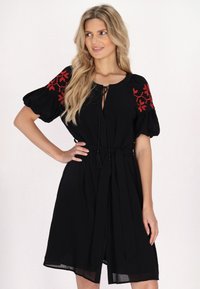 Schwarzes Kleid mit roten bestickten Akzenten an den Puffärmeln, einem Bindegürtel in der Taille und einem V-Ausschnitt. Leichter Stoff, knielanges Design.