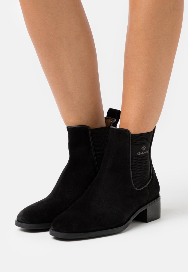 Bottes cheville en daim noir avec talons blocs bas, portées sur jambes nues, dotées de panneaux latéraux élastiques et de languettes à l'arrière.