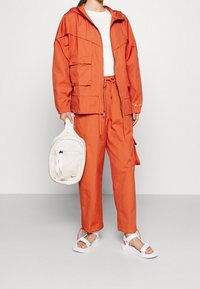 Orange lättviktsjacka med huva, två framfickor, matchande byxor och vita sandaler som håller en gräddfärgad ryggsäck med dragkedja.