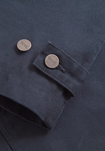 Manșeta cămășii din țesătură albastru închis, cu două nasturi argintii, care au gravat „W.W.”. Textura este ușor aspră, evidențiind designul său durabil.