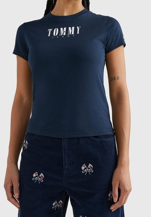Kobieta w granatowym krótkim rękawie koszulce „Tommy Jeans” i granatowych spodniach sztruksowych z małymi haftowanymi flagami.