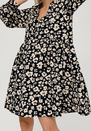 Kvinde iført en sort knælængde kjole med beige blomsterprint og lange ærmer, løfter armene lidt over hovedet.