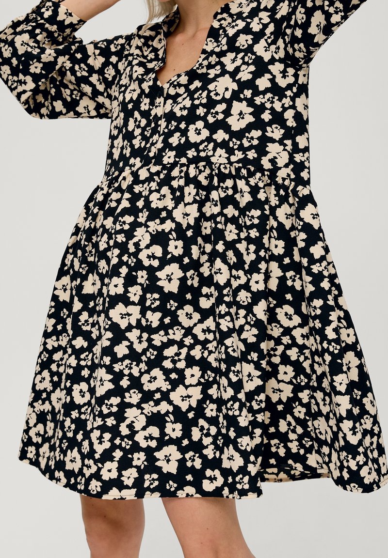 Femme portant une robe noire mi-longue avec imprimé floral beige et manches longues, levant les bras légèrement au-dessus de la tête.