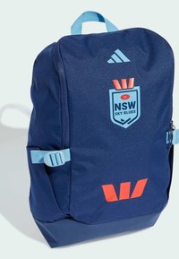 Zaino blu navy realizzato in tessuto resistente, con un logo scudo prominente, spallacci regolabili e dettagli rossi sulla parte anteriore.
