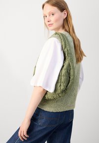 Groene puffer vest met pluizige textuur, gedragen over een witte blouse met opbollende mouwen; gecombineerd met donkerdenim jeans.
