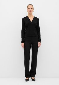 Top nero a maniche lunghe con scollo a V e dettagli plissettati, abbinato a pantaloni neri aderenti e scarpe nere basse. Design semplice ed elegante.