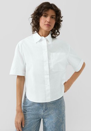 Kobieta o kręconych włosach, ubrana w białą koszulę z krótkim rękawem zapinaną na guziki oraz jasnoniebieskie dżinsy w kwiatowy wzór, stojąca na tle jednolitego tła.