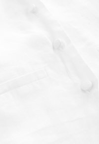 Fehér lenvászon ing, sima textúrával, négy hozzáillő anyaggombbal és egy kis zsebdísszel a bal oldalon.