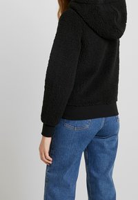 Czarny polarowy hoodie o teksturowej powierzchni, w oversize'owym fasonie, z kapturem. W zestawie z jasnoniebieskimi szerokimi spodniami dżinsowymi.