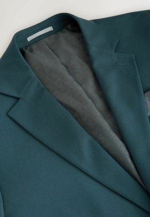 Teal bleiseri, jossa on teksturoitu pinta ja sileä harmaa kuvioitu vuori. Huomiota herättäviä yksityiskohtia ovat läpän kaulus ja yksivetoinen kiinnitys.