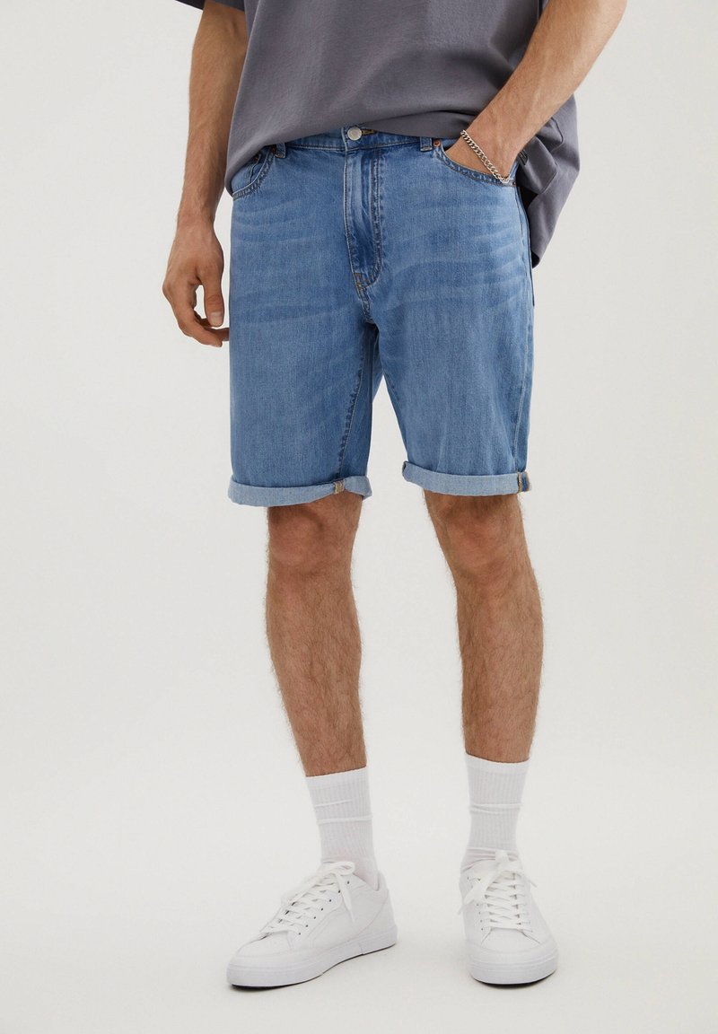 Shorts en denim bleu clair avec des revers, poches avant et passants de ceinture ; associés à des baskets blanches et des chaussettes de cheville.