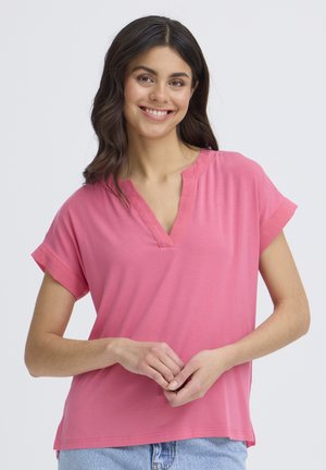 Jonge vrouw met donker haar lacht, draagt een roze T-shirt met korte mouwen en V-hals en lichtblauwe spijkerbroek tegen een effen achtergrond.