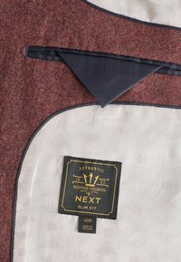 Tissu tweed bordeaux avec un revers bleu marine, doublure à motifs crème et étiquette indiquant "Authentic Heritage Tailoring NEXT Slim Fit, 40R, fabriqué au Vietnam."