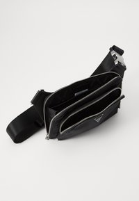 Bolso cruzado de nylon negro con tres compartimentos con cremallera y una correa trenzada, con herrajes plateados y un detalle de logo discreto.