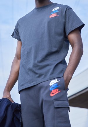 Man met een donkergrijs Nike T-shirt en broek met witte, blauwe en rode Nike-logo's, staand met één hand in de zak en een jas vasthoudend.