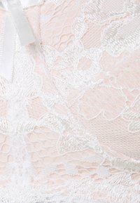 Bralette rendado em rosa suave com padrões florais brancos intrincados. Apresenta um brilho delicado e um detalhe de laço em cetim. Camada de renda texturizada.